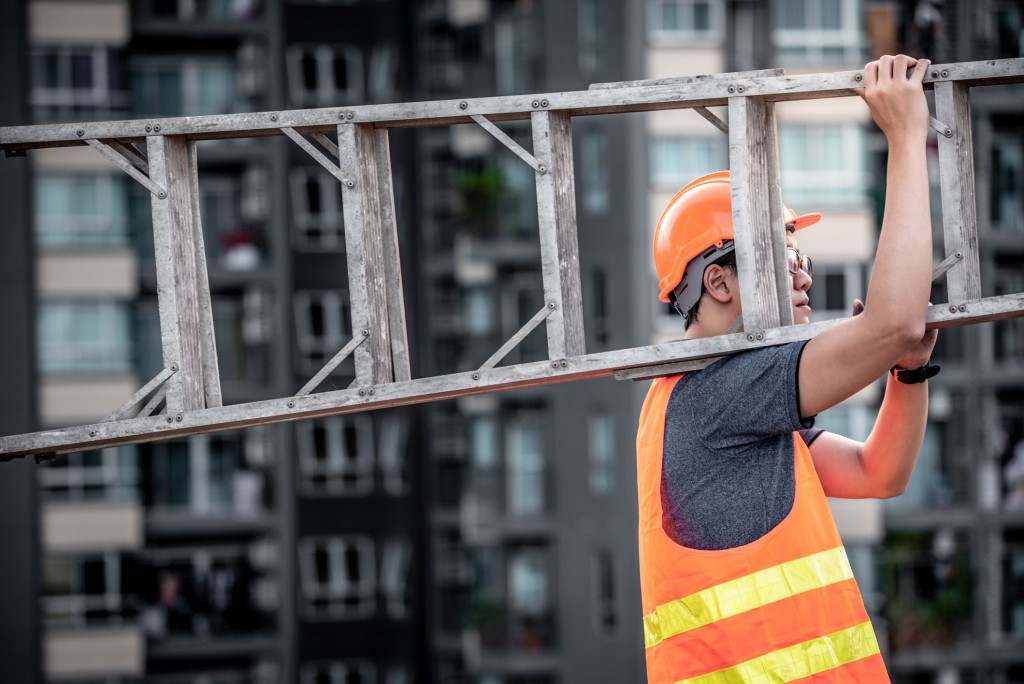 Maintenance worker with orange safety helmet and vest carrying aluminium step ladder at construction site. 